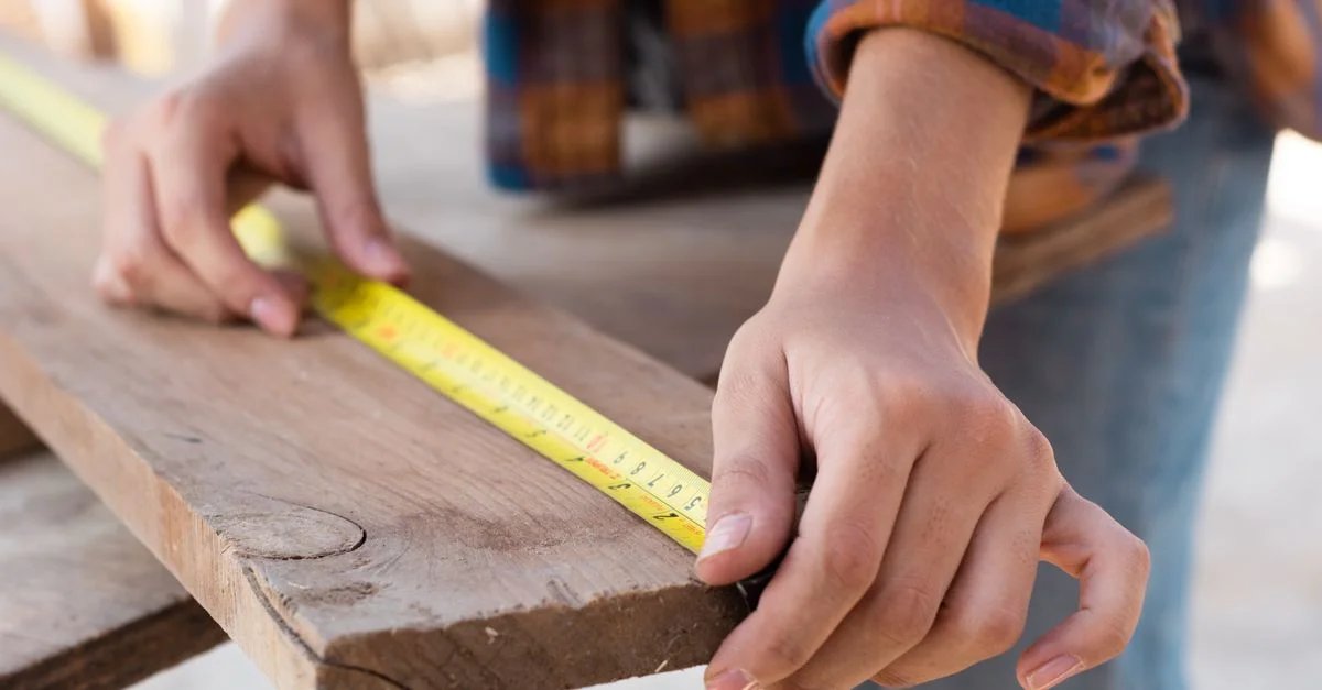 Gros plan des mains mesurant une planche en bois avec un mètre ruban dans un contexte de menuiserie.