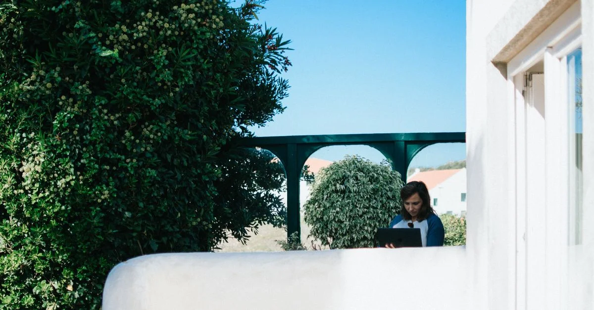 Une femme travaillant sur son ordinateur portable dans un jardin ensoleillé avec une végétation luxuriante et un ciel bleu clair.
