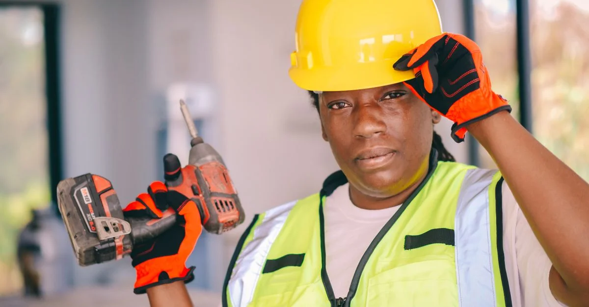 Femme de construction confiante portant un équipement de sécurité et tenant une perceuse à l'intérieur.