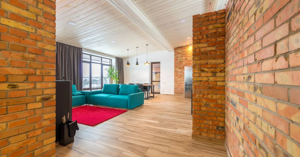 Salon spacieux avec des murs en briques, un canapé teal et de grandes fenêtres laissant entrer la lumière naturelle.