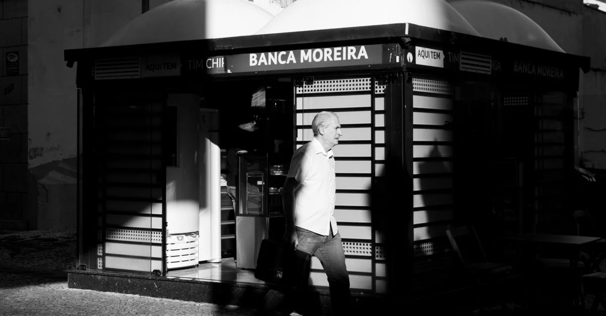 Photo de rue en noir et blanc d'un homme âgé marchant devant le kiosque Banca Moreira à Curitiba, au Brésil.