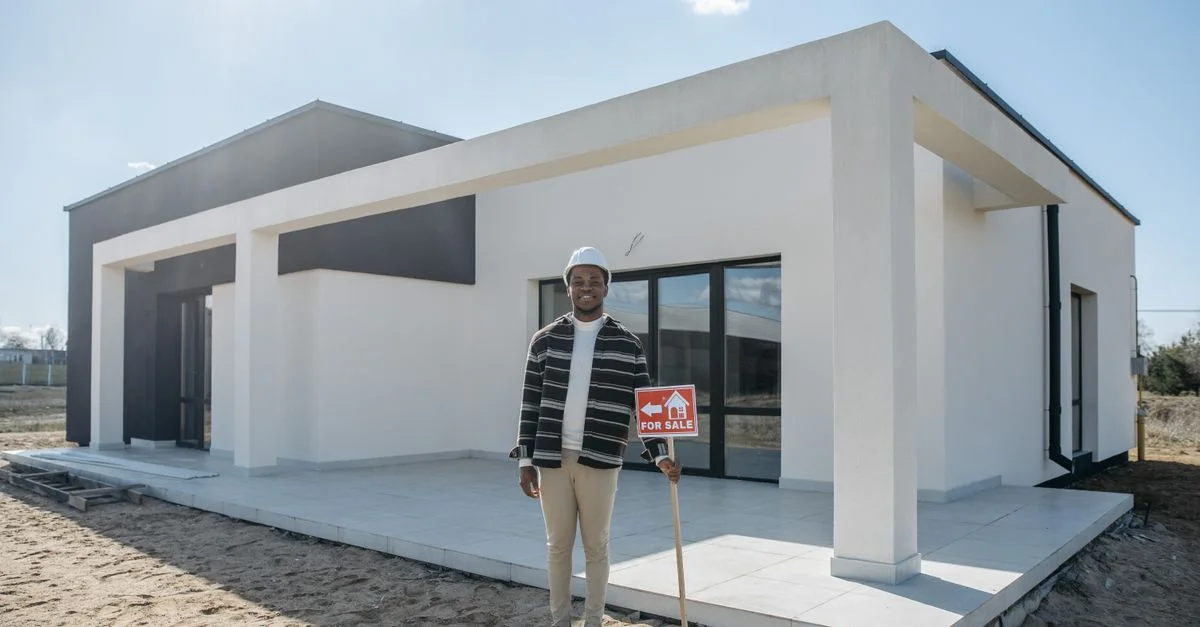 Un agent immobilier avec un casque de protection tient un panneau À vendre devant une maison moderne, prêt à être vendu.